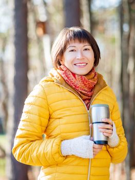 Happy wide smiling women in bright yellow jacket is holding thermos mug. Hot tea or other beverage on cool autumn day.