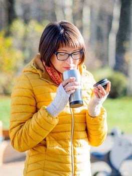 Happy wide smiling women in bright yellow jacket is holding thermos mug. Hot tea or other beverage on cool autumn day.