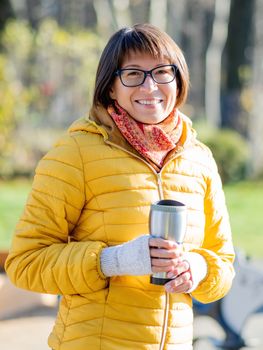 Happy wide smiling women in bright yellow jacketis holding thermos mug. Hot tea or other beverage on cool autumn day.