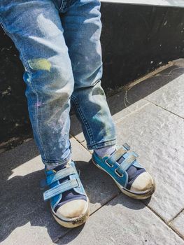 Toddler in jeans draws with crayons on the asphalt in sunny day. Kid's jeans and sneakers are covered with colorful stains. Outdoor leisure activity.