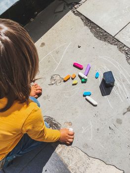 Toddler in jeans draws with crayons on the asphalt in sunny day. Child is holding colored crayons. Kid's hands and clothes are covered with colorful chalks. Outdoor leisure activity.