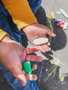 Toddler in jeans draws with crayons on the asphalt in sunny day. Child is holding colored crayons. Kid's hands and clothes are covered with colorful chalks. Outdoor leisure activity.