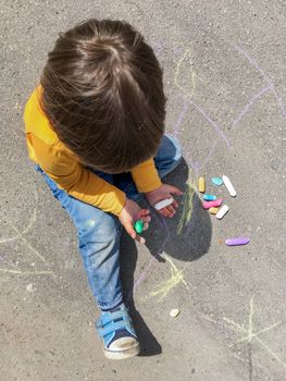 Toddler in jeans draws with crayons on the asphalt in sunny day. Child is holding colored crayons. Kid's hands and clothes are covered with colorful chalks. Outdoor leisure activity.