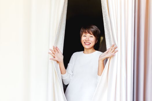 Wide smiling beautiful asian woman opens curtains on window. Young happy woman among textile folds of drapery.