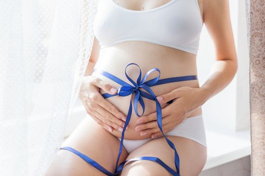 Pregnant woman in white underwear with blue bow on belly. Young woman expecting a baby boy. Cozy happy background in sunny morning.