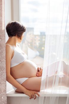 Pregnant woman in white underwear sitting on window sill. Young woman expecting a baby. Cozy happy background in sunny morning.