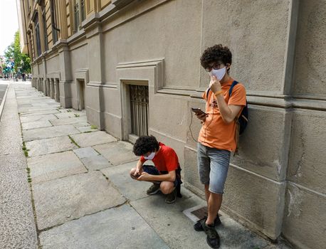 Two Caucasian boys use smartphones in the time of the coronavirus: one crouched and the other leaning against the wall. They wear a white protective mask.