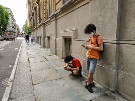 Two Caucasian boys use smartphones in the time of the coronavirus: one crouched and the other leaning against the wall. They wear a white protective mask.