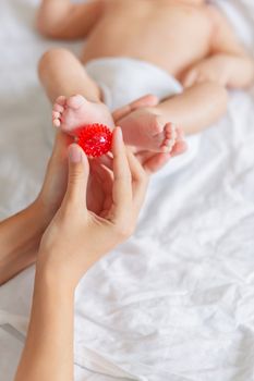 Mother holds newborn baby's feet. Tiny fingers and red massage ball in woman's hand. Cozy morning at home.