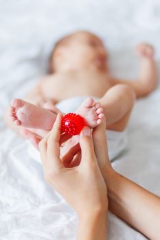 Mother holds newborn baby's feet. Tiny fingers and red massage ball in woman's hand. Cozy morning at home.