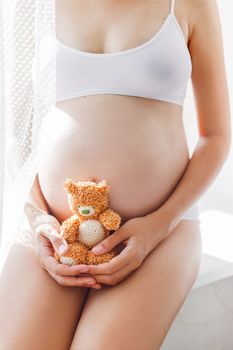 Pregnant woman in white underwear with knitted toy teddy bear. Young woman expecting a baby. Cozy happy background in sunny morning.