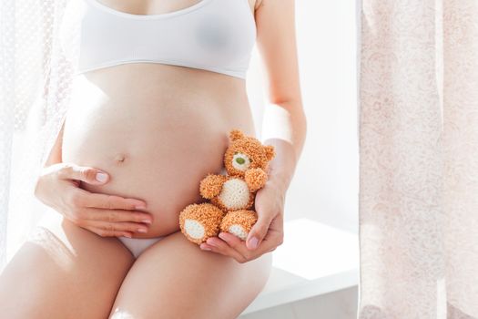 Pregnant woman in white underwear with knitted toy teddy bear. Young woman expecting a baby. Cozy happy background in sunny morning.