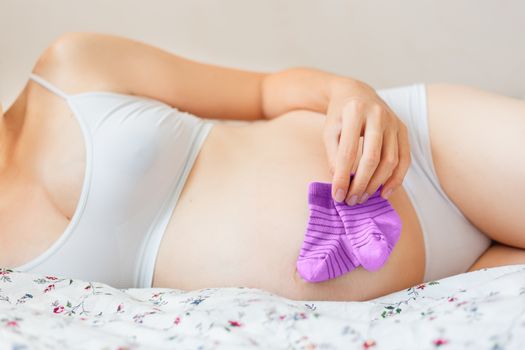 Pregnant woman in white underwear lying in bed with small baby's socks. Young woman expecting a baby girl.