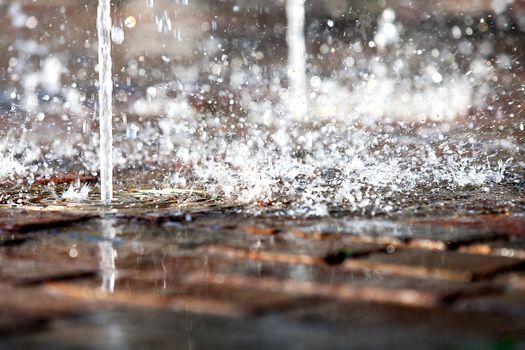 The stream of water hits into a paved pavement. Splashes of water sparkle on the sun.