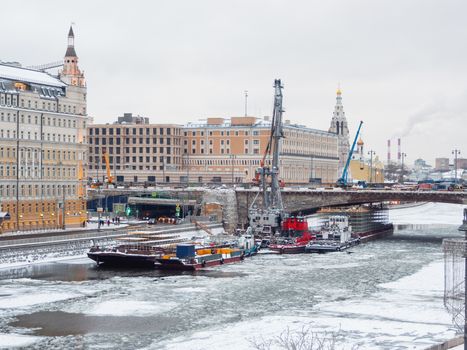 Ships with construction equipment are in Moscow-river under the bridge. Reconstruction of Bolshoy Moskvoretsky Bridge. Winter cloudy day in Moscow, Russia.