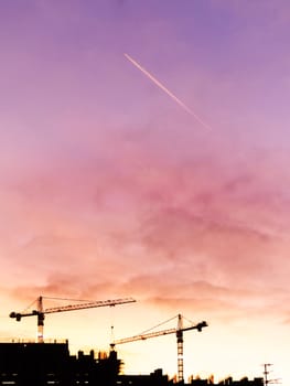 Gorgeous pink, coral and violet sunset with clouds, flying airplane and construction cranes. Colorful sky upon construction site.