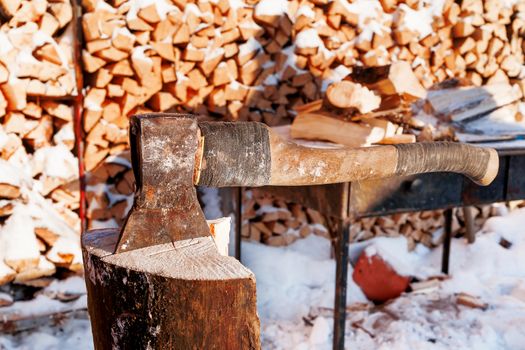 Axe in a wooden deck. Winter sunny day. Rural background with folded pile of firewood.