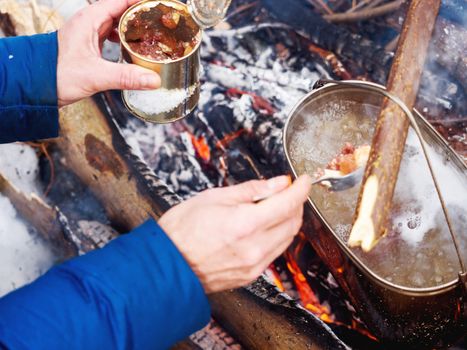 Cooking soup on a fire pot. Tourist puts stewed meat in soup from tin can. Winter camping in forest.