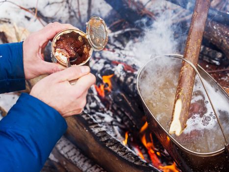 Cooking soup on a fire pot. Tourist puts stewed meat in soup from tin can. Winter camping in forest.