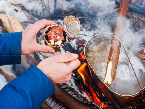 Cooking soup on a fire pot. Tourist puts stewed meat in soup from tin can. Winter camping in forest.