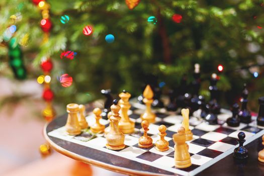 Wooden chess set on the table under decorated Christmas tree. Quiet game holiday winter evening. Family leisure.