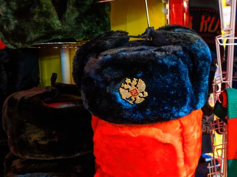 Colorful Russian winter hats with earflaps. Metal pin with Russian double-headed eagle symbol. Souvenir shop in Moscow, Russia.