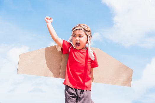 Happy Asian funny child or kid little boy smile wear pilot hat and goggles play toy cardboard airplane wing flying raises hand up against summer blue sky cloud background, Startup freedom concept