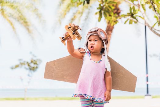 Happy Asian funny child or kid little girl smile wear pilot hat and goggles play toy cardboard airplane wing flying against summer sky cloud on trees garden background, Startup freedom concept