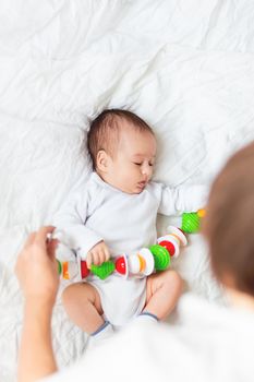 Woman plays with newborn boy. Baby's first toy - colorful rattle garland. Top view on little child in white onesie. Morning bedtime in cozy home.