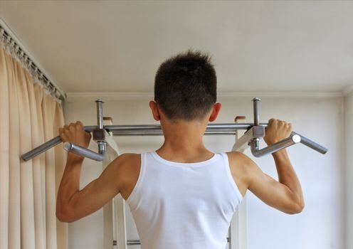 A teenager in a white T-shirt does strength exercises on the crossbar in the home sports corner.