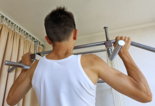 A teenager in a white T-shirt does strength exercises on the crossbar in the home sports corner.