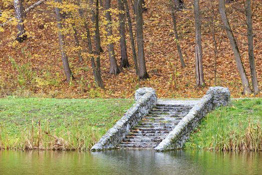 On the shore of the autumn forest pond, the old, moss-covered, stone steps touch the calm surface of the water.