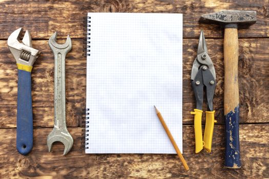 Old hand tools, hammer, metal shears, adjustable wrench, carob wrench surround an empty notebook with a pencil against the background of an old wooden table.