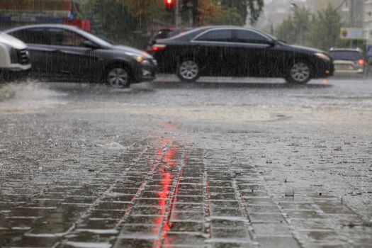 Torrential rain on the road and on the sidewalk pours on passing cars illuminated by a red traffic light.