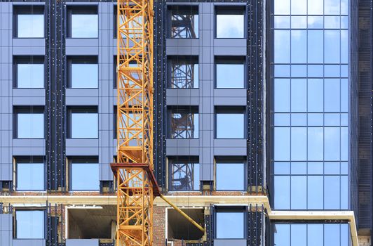 A fragment of the facade of a house under construction, a modern building is being built, a tower crane and a blue sky are reflected in the glass walls of the building.