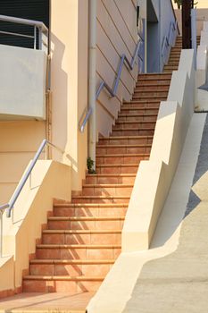 The path goes up a stone staircase from beige paving slabs in the bright sun in a narrow Greek street.