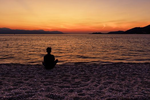 A silhouette of a teenager who sits on a beach of small pebbles of the seashore and watches a beautiful, bright sunset on the Corinthian Gulf, Loutraki, Greece,