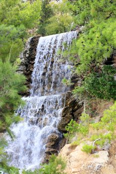 A large waterfall from a spring of radon water flows down large boulders at the foot of the mountain among the green Mediterranean pines and palm trees in the park of Loutraki, Greece, vertical image.
