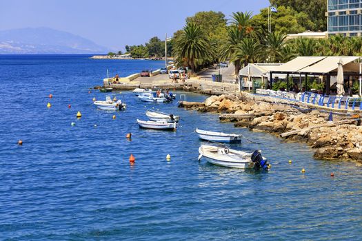 Powerboats and boats are moored at the Gulf of the Ionian Sea against the backdrop of the sunlit promenade, palm alley along the city road and mountain range.