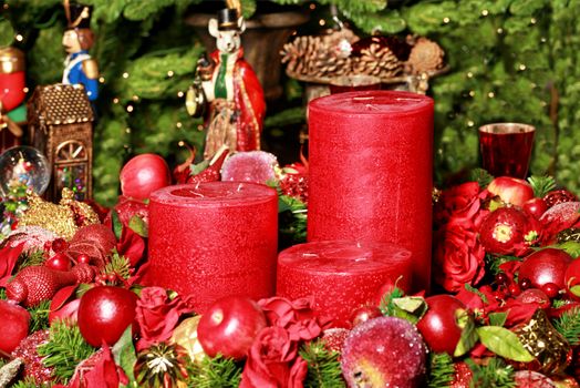 Christmas decoration: three thick red candles surrounded by blurry Christmas decorations and fairytale figures on the background of fir branches, close-up.