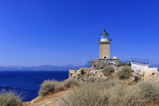 An old stone lighthouse stands on the high cape of Malagavi against the backdrop of the blue sea and sky in the afternoon sun, Loutraki, Greece.