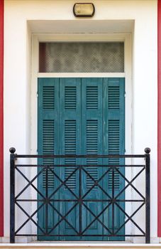 Old wooden balcony doors with wooden shutters with metal grilles of the balcony, framed on the facade along the perimeter with red vertical stripes.