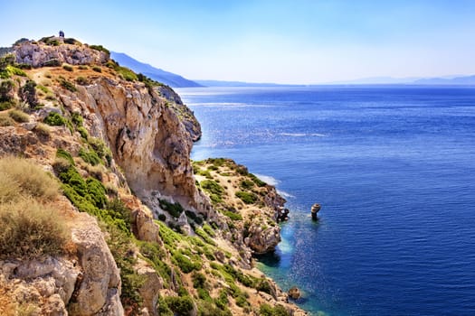 Calm turquoise waves wash the rocky shore of the Gulf of Corinth in the Ionian Sea in the rays of the dazzling and hot sun.