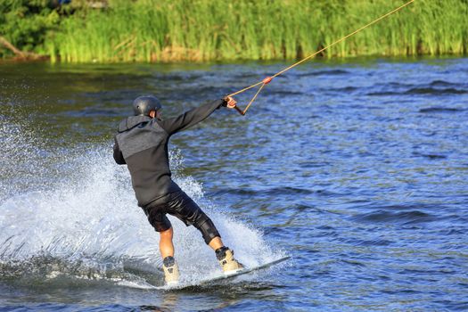 A wakeboarder rushes through the water at high speed along the banks of the river, holding the cable pulled with one hand and creating a cloud of spray around hiself.