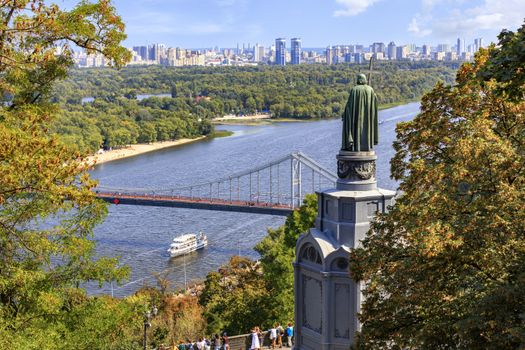 Autumn cityscape of Kyiv overlooking the pedestrian bridge to Trukhanovy island, the monument to Volodymir and the blue waters of the Dnipro River on which pleasure boats go.