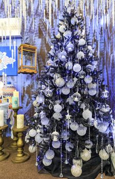 A collection of silver stars, Christmas balls, Christmas baubles, decorative icicles is illuminated by blue lights on a slender Christmas tree.
