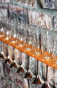 Glass champagne glasses lined up on a shelf diagonally near an old stone wall, whith copyspace for text.