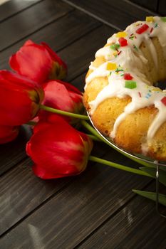 Easter yeast cake or babka covered with icing and decorated with colorful decoration on a dark wooden table and bright tulips, vertical image