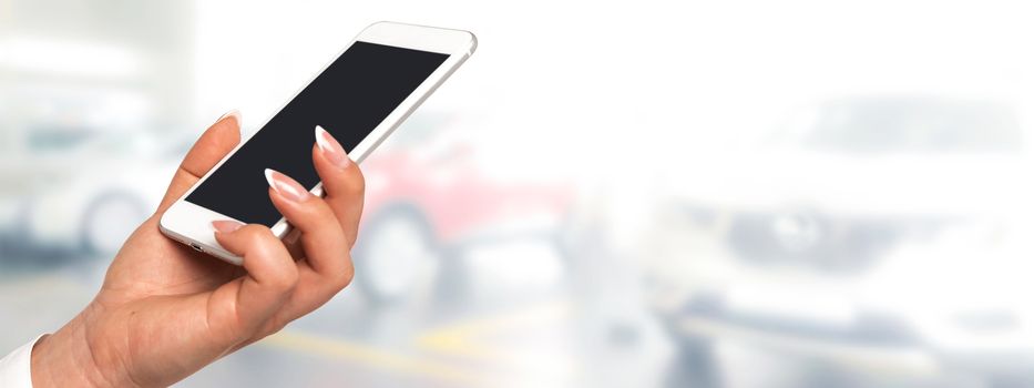 Smart car concept banner - female hand  holding smartphone with blank screen on a blurred background of the auto dealership (copy space).