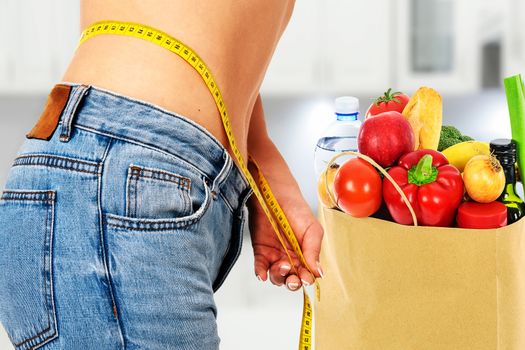 Healthy eating concept - woman in blue jeans measures her perfect waistline with a measuring tape next to a bag of dietetic food in the kitchen (mixed).